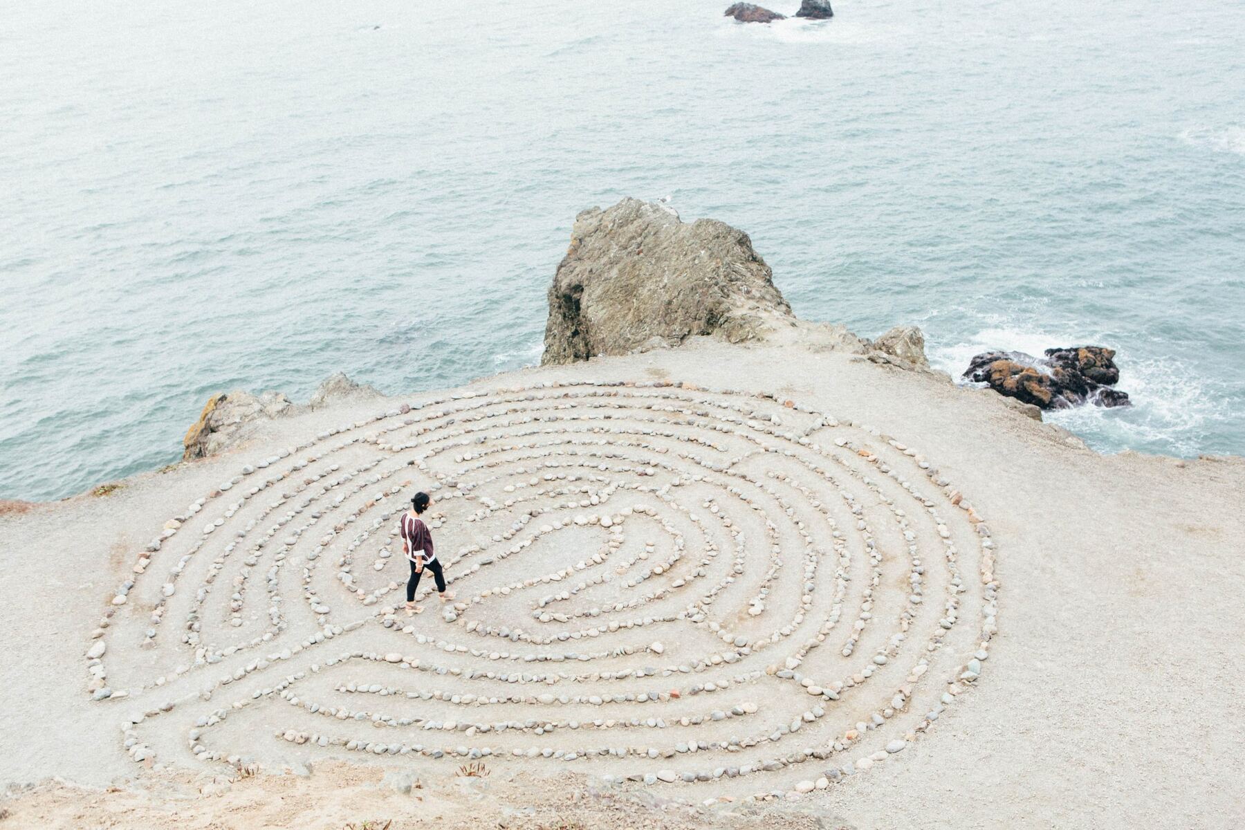 Eine Frau geht barfuß durch ein Steinlabyrinth auf einer Klippe über dem Meer.
